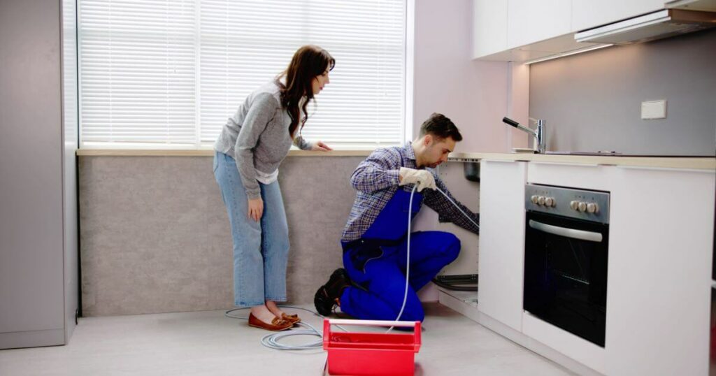 Emergency plumber in blue overalls fixing a kitchen sink while a woman watches attentively, showcasing prompt plumbing services.